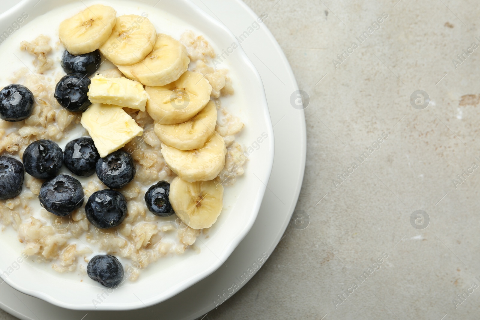 Photo of Tasty oatmeal with banana, blueberries, butter and milk served in bowl on light grey table, top view. Space for text