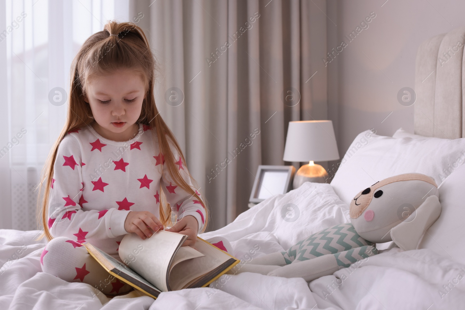 Photo of Cute little girl reading book on bed at home