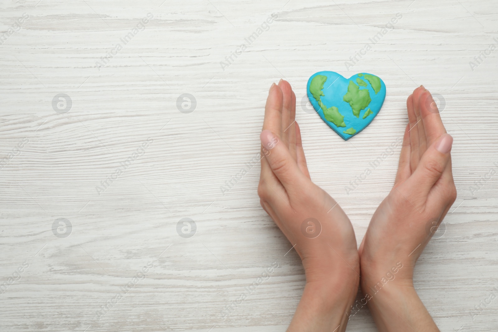 Photo of Happy Earth Day. Woman with heart shaped plasticine planet at white wooden table, top view and space for text