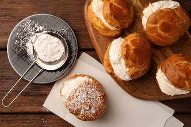 Delicious profiteroles with cream filling and powdered sugar on wooden table, flat lay