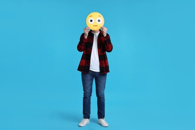 Man covering face with surprised emoticon on light blue background