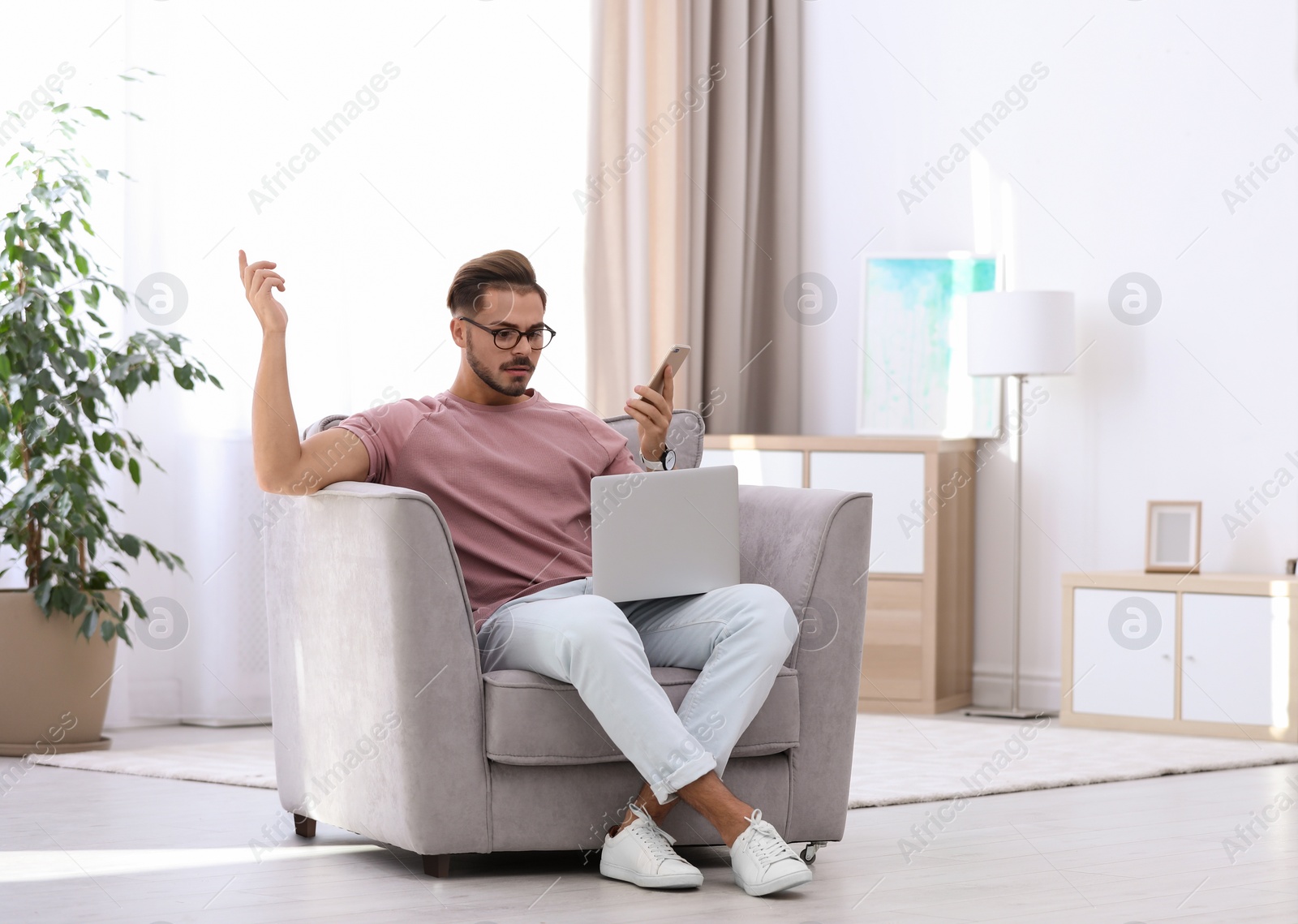 Photo of Emotional young man with laptop and smartphone in armchair at home. Space for text