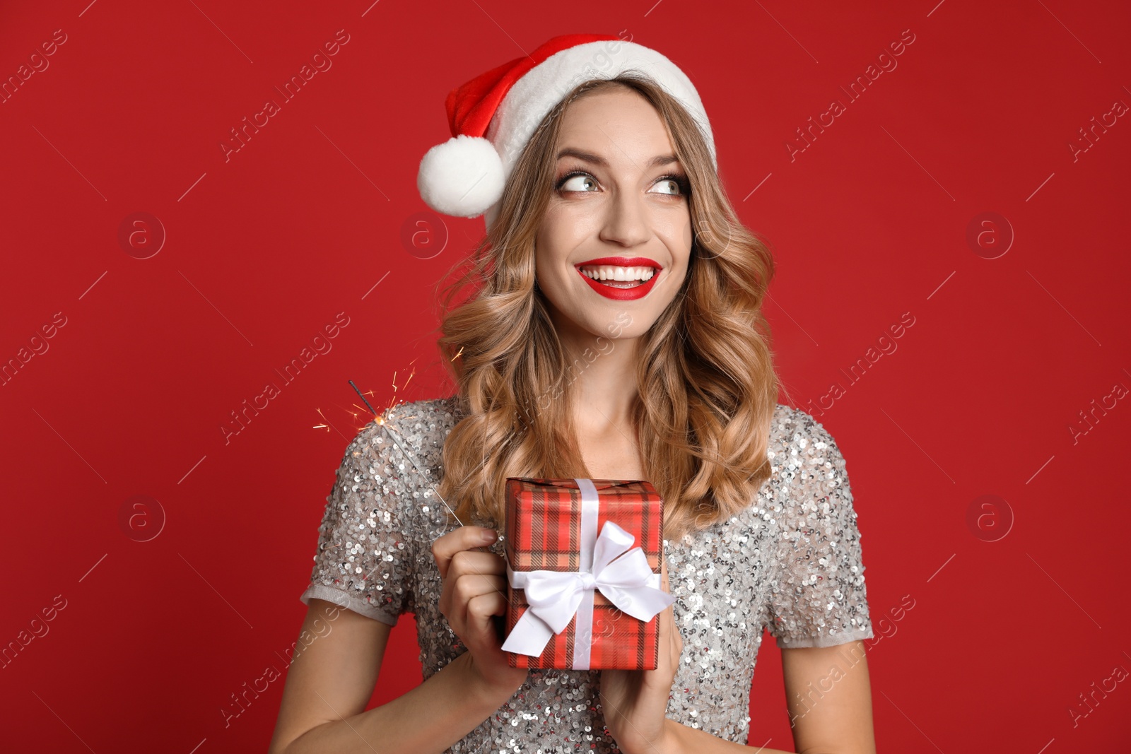 Photo of Happy young woman wearing Santa hat with Christmas gift and sparkler on red background