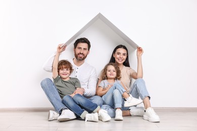 Photo of Family housing concept. Happy woman and her husband holding plastic roof while sitting with kids on floor at home