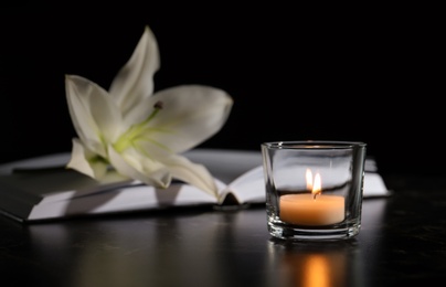 Burning candle, book and white lily on table in darkness, space for text. Funeral symbol