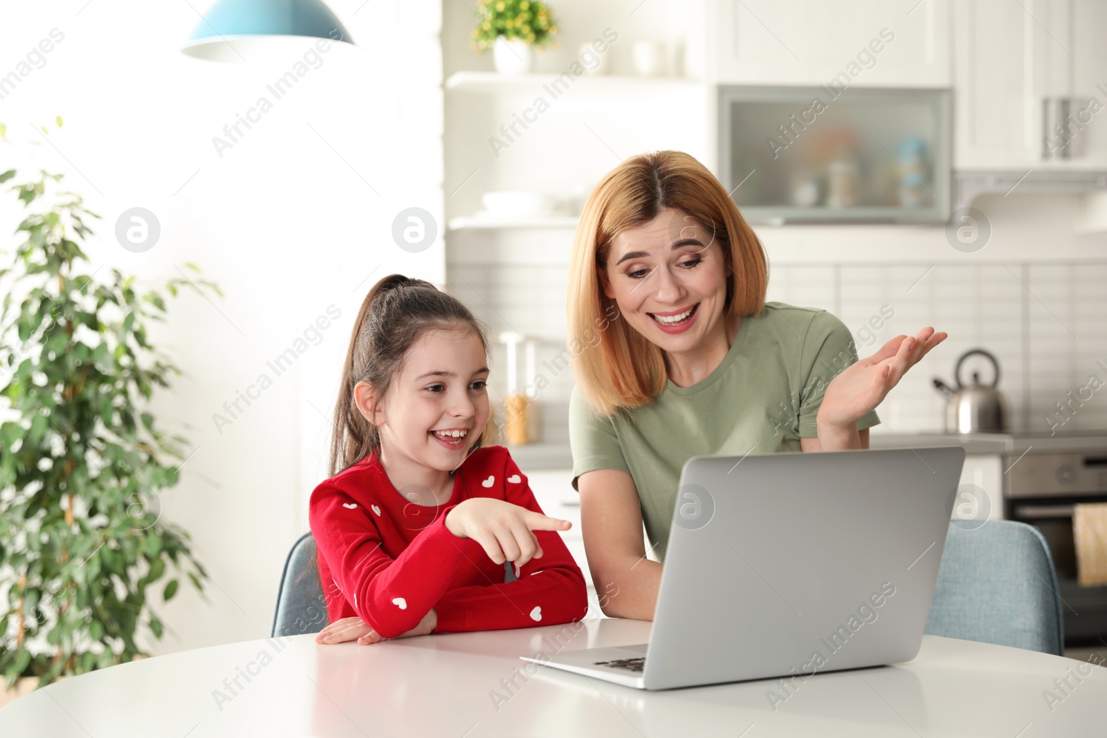 Photo of Mother and her daughter using video chat on laptop at table in kitchen