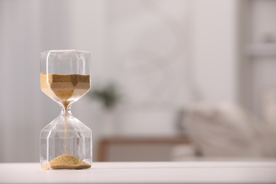 Photo of Hourglass with flowing sand on white table against blurred background. Space for text