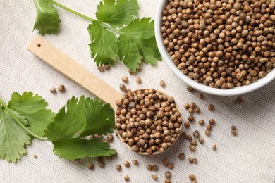 Dried coriander seeds in bowl, spoon and green leaves on light cloth, flat lay