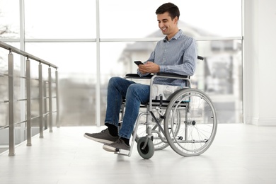Young man in wheelchair using mobile phone near window indoors