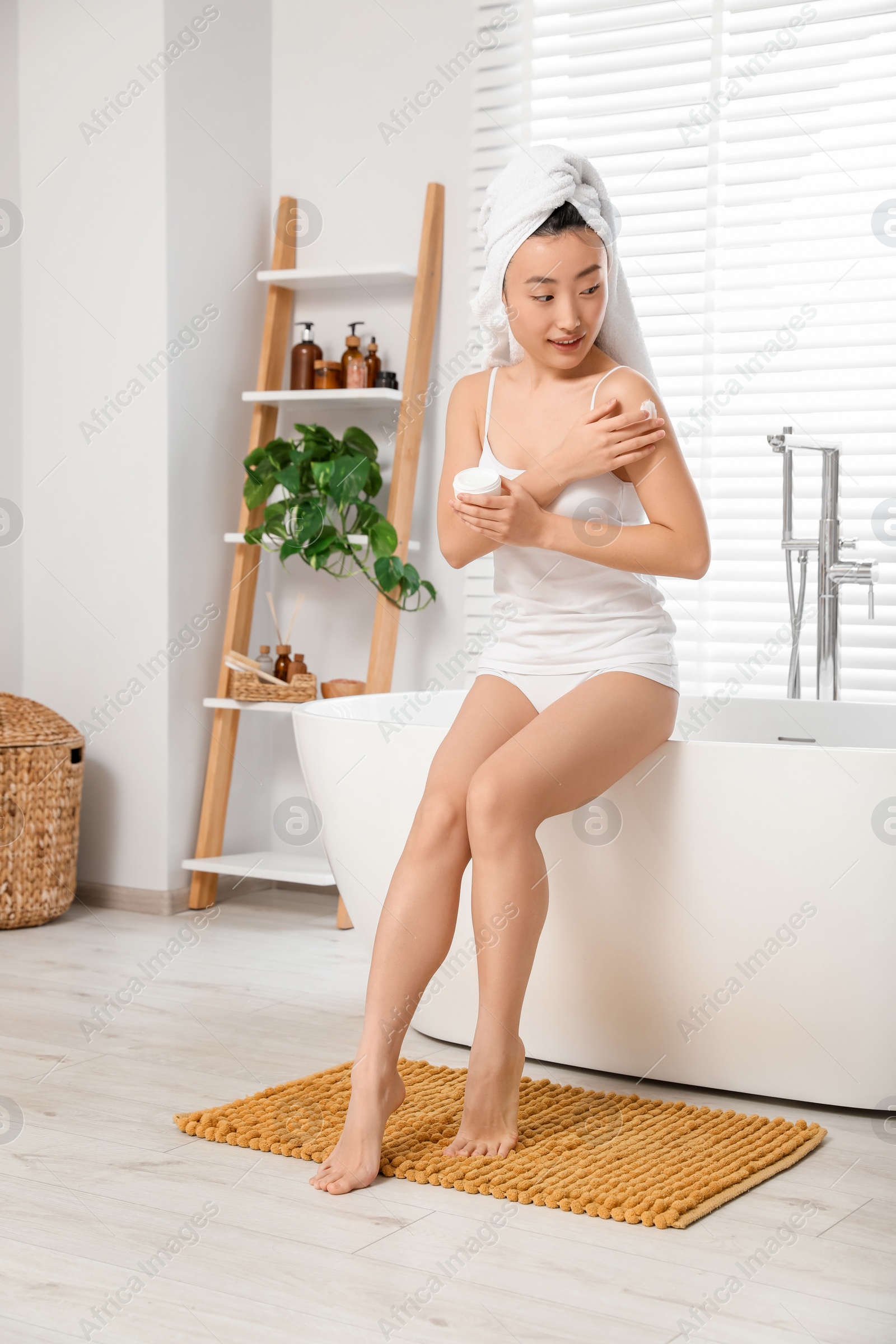 Photo of Beautiful young Asian woman applying body cream on shoulder in bathroom, space for text