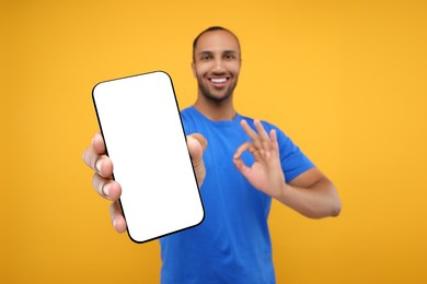 Photo of Young man showing smartphone in hand and OK gesture on yellow background, selective focus. Mockup for design