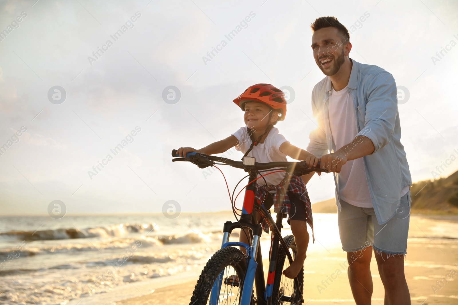 Photo of Happy father teaching son to ride bicycle on sandy beach near sea at sunset