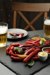 Photo of Different thin dry smoked sausages, sauces and glasses of beer on grey table