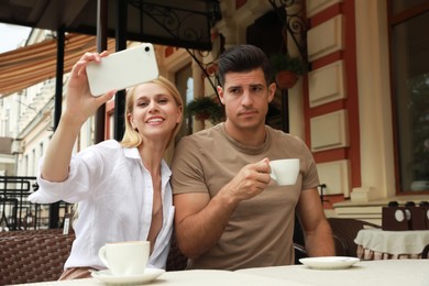 Photo of Young woman trying to take selfie with her displeased boyfriend in outdoor cafe. Boring date