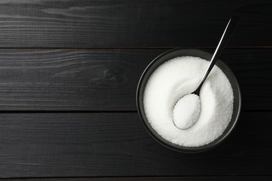 Photo of Granulated sugar in bowl and spoon on black wooden table, top view. Space for text