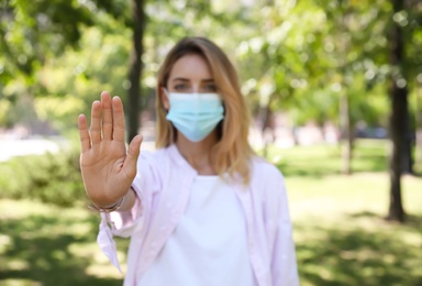 Woman in protective face mask showing stop gesture in park, focus on hand. Prevent spreading of coronavirus