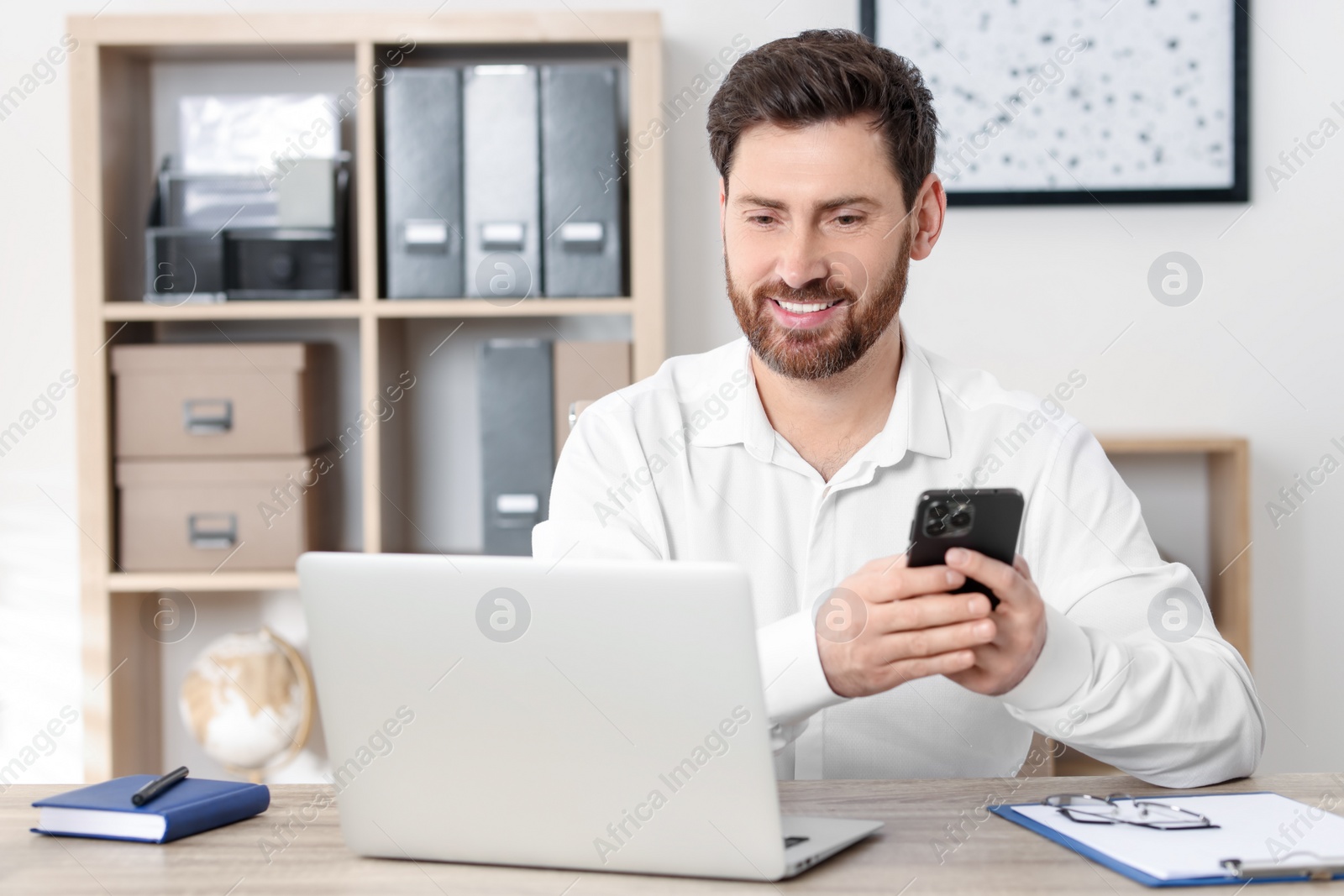 Photo of Smiling man with smartphone using laptop at table in office