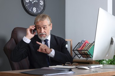 Emotional senior boss talking on phone at wooden table in modern office