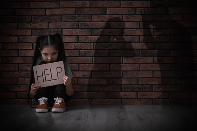 Sad little girl with sign HELP sitting on floor and silhouettes of arguing parents 