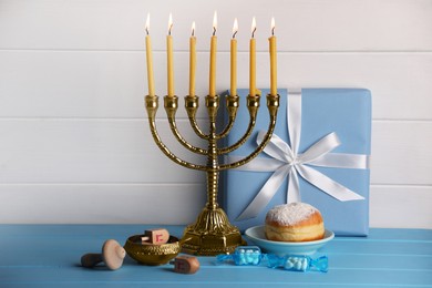 Photo of Hanukkah celebration. Menorah with burning candles, dreidels, donut and gift box on light blue wooden table