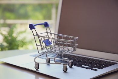 Photo of Internet shopping. Laptop with small cart on table indoors, closeup