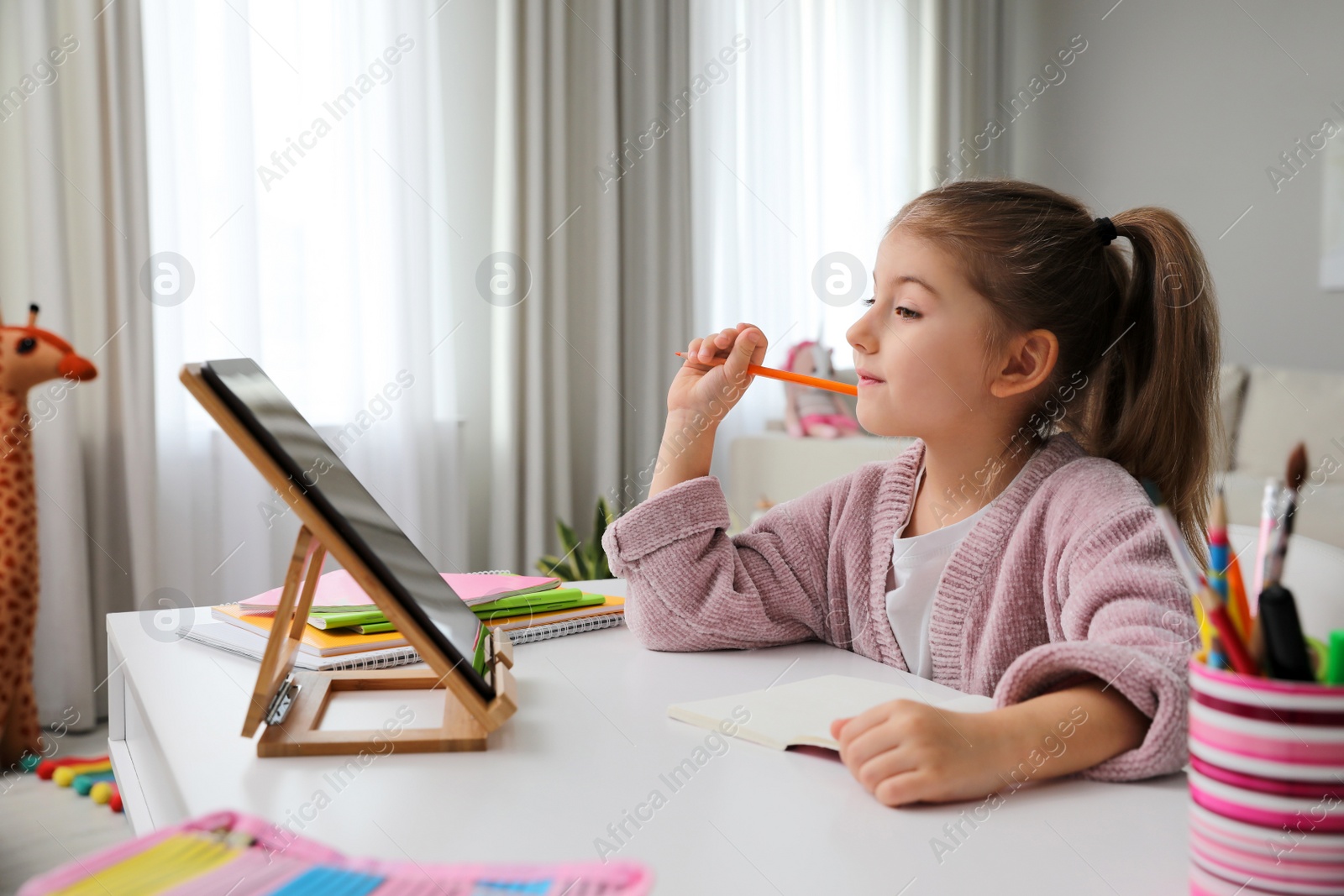 Photo of Adorable little girl doing homework with tablet at table indoors