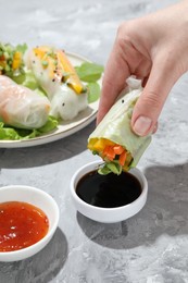 Photo of Woman dipping delicious spring roll into soy sauce at grey textured table, closeup