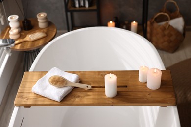 Photo of Wooden tray with burning candles, towel and brush on bathtub in bathroom