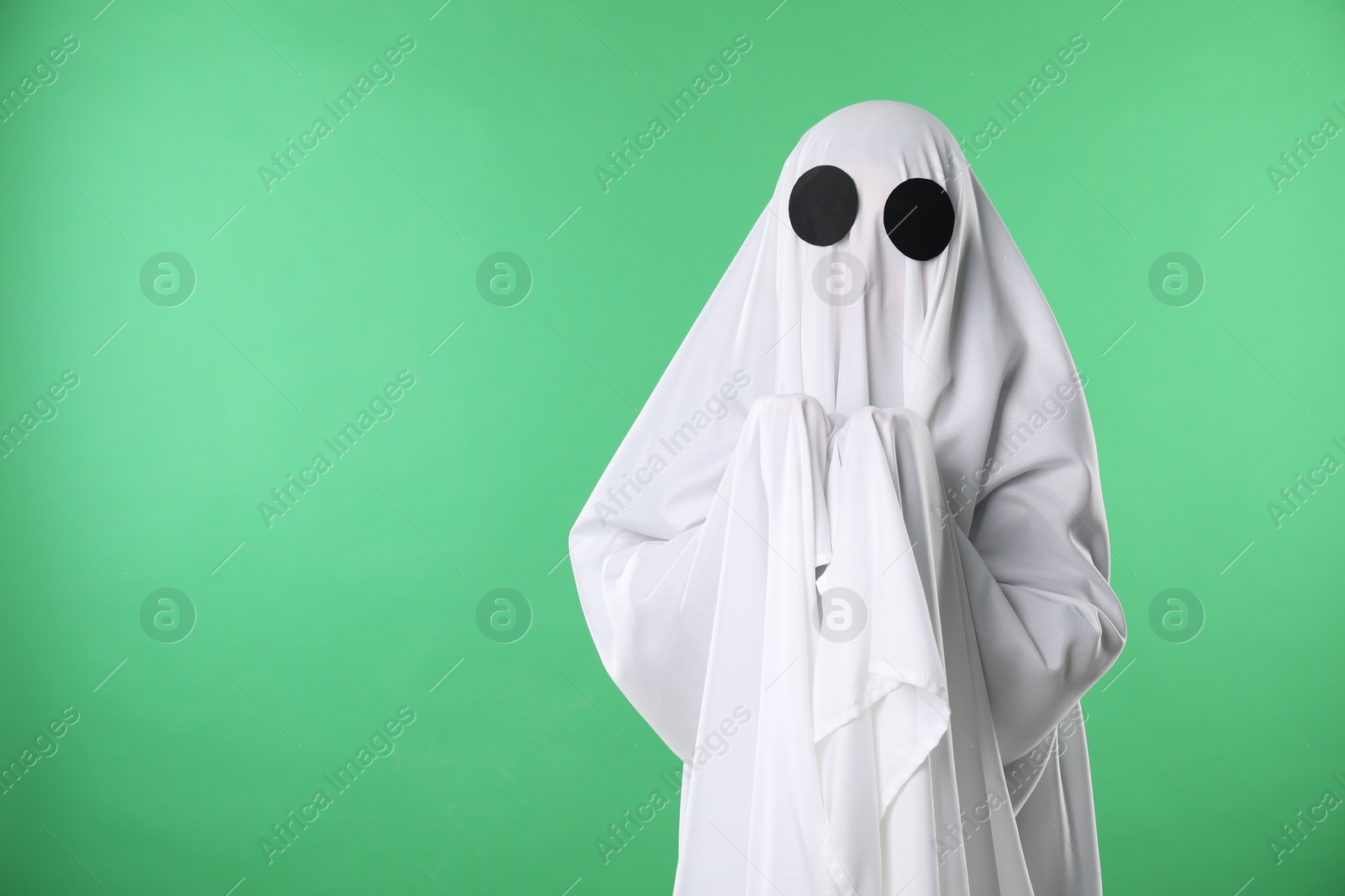 Photo of Creepy ghost. Person covered with white sheet on green background, space for text