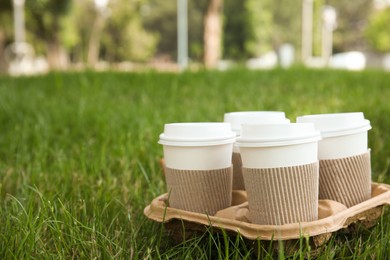 Photo of Takeaway paper coffee cups with plastic lids and sleeves in cardboard holder on green grass outdoors, space for text