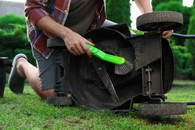 Photo of Man cleaning lawn mower with brush in garden, closeup