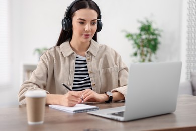 Photo of Young woman in headphones watching webinar at table in room