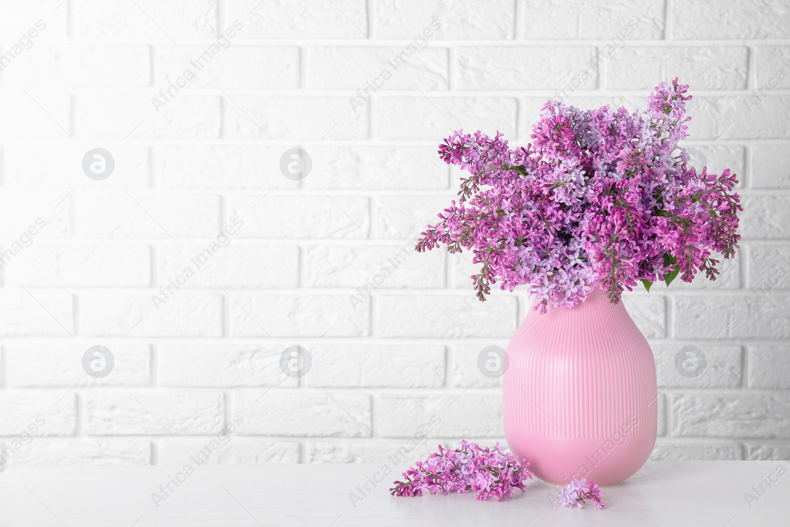 Photo of Blossoming lilac flowers in vase on light table against brick wall. Space for text