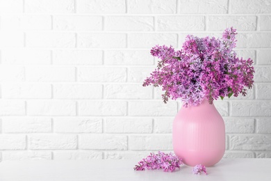 Photo of Blossoming lilac flowers in vase on light table against brick wall. Space for text