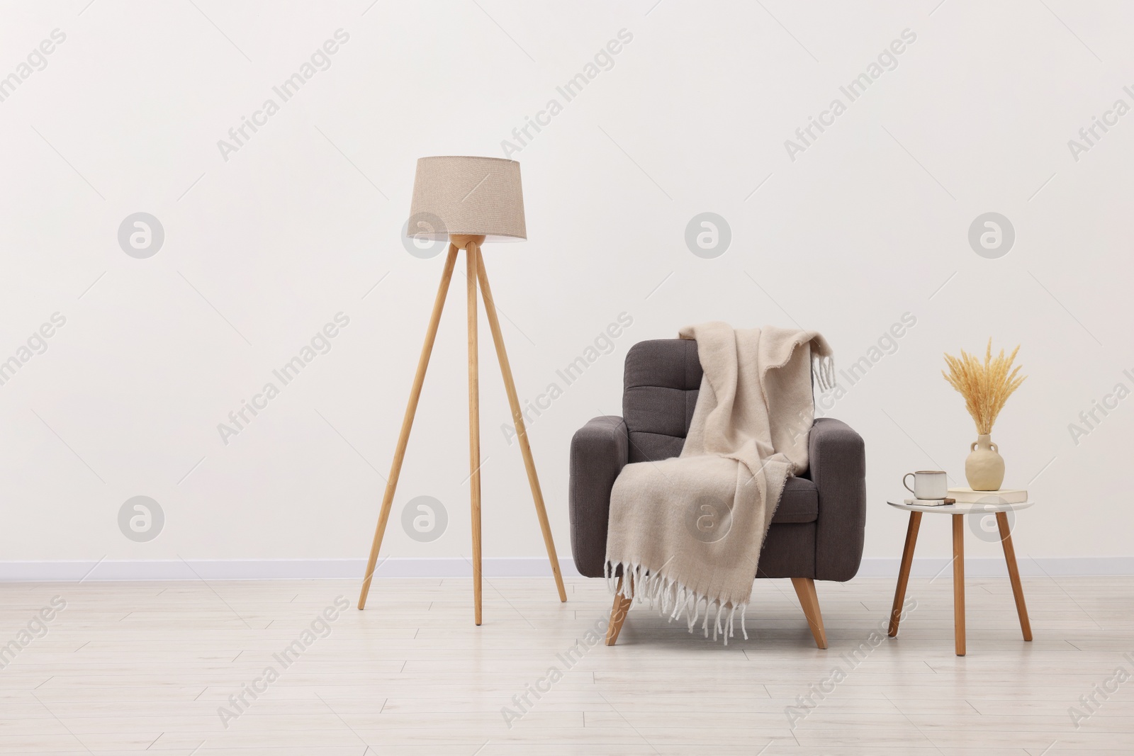 Photo of Comfortable armchair, blanket, lamp and side table near white wall indoors