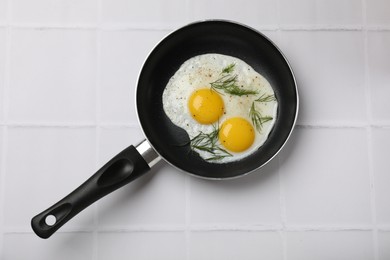 Photo of Frying pan with tasty cooked eggs and dill on white tiled table, top view