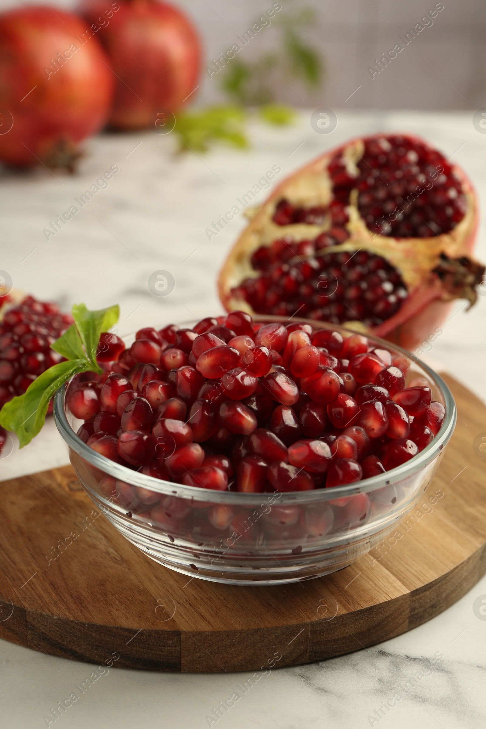 Photo of Ripe juicy pomegranate grains in bowl and green leaves on white marble table