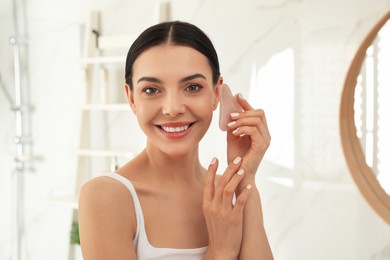 Photo of Beautiful young woman doing facial massage with gua sha tool in bathroom