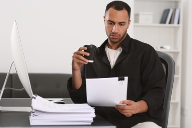 Photo of Man drinking coffee while working with documents at table in office