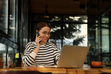 Young female student with laptop talking on phone while studying at table in cafe