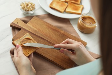 Photo of Woman spreading tasty nut butter onto toast at white table, closeup