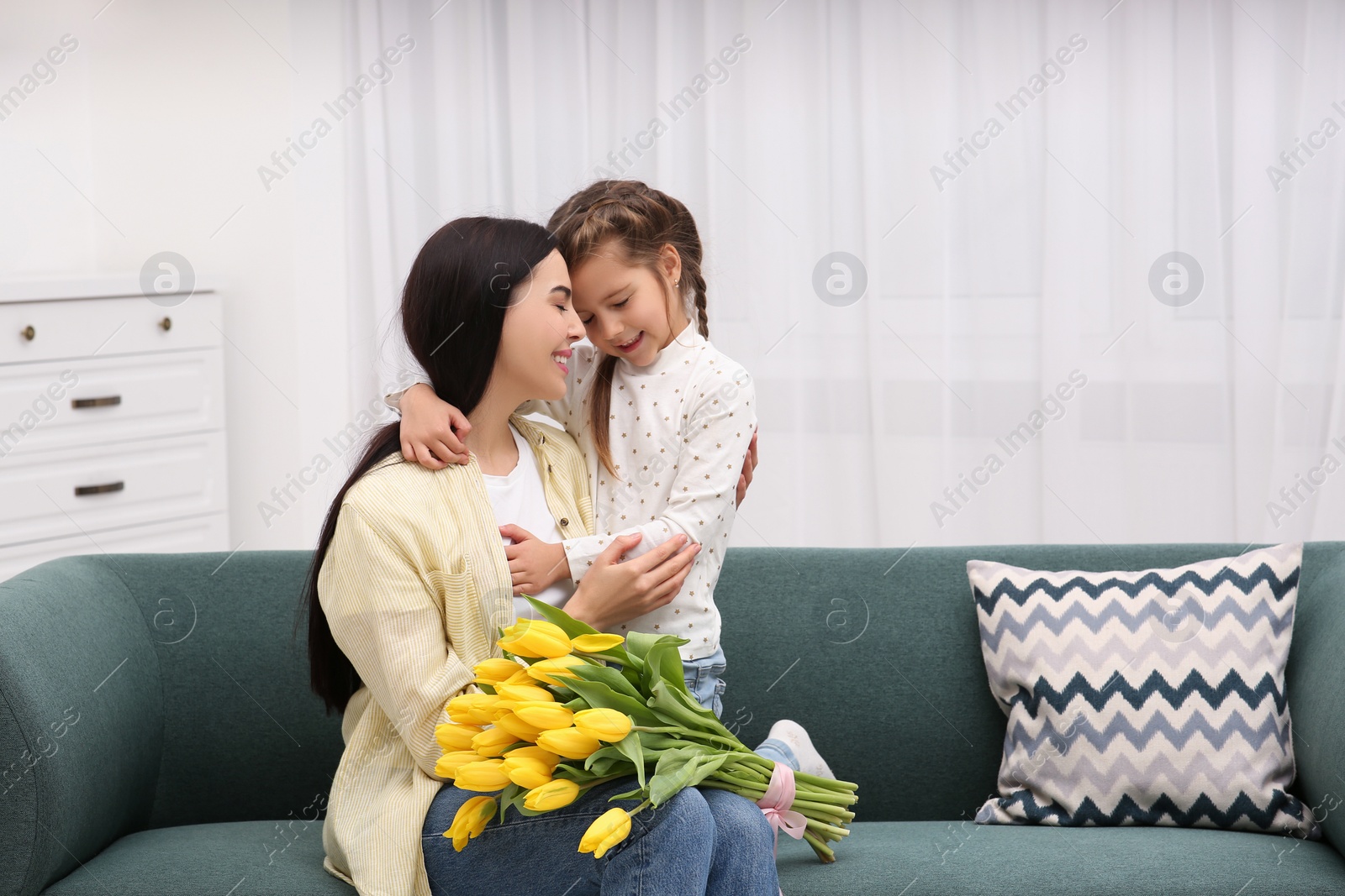 Photo of Happy woman with her daughter and bouquet of yellow tulips on sofa at home, space for text. Mother's day celebration