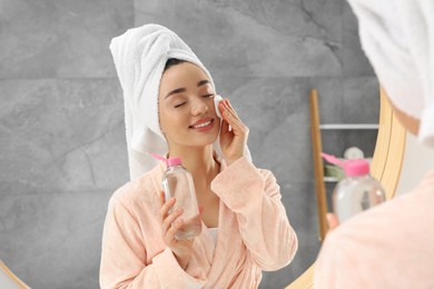 Photo of Beautiful woman in terry towel removing makeup with cotton pad near mirror indoors