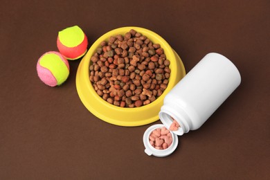 Bowl with dry pet food, bottle of vitamins and toys on brown background, above view