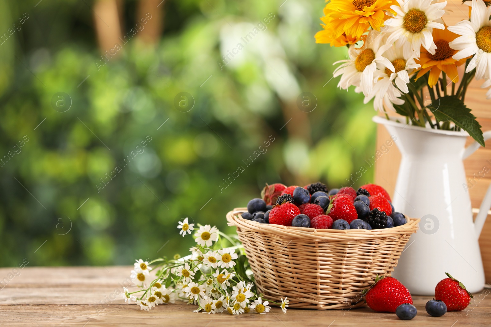 Photo of Wicker bowl with different fresh ripe berries and beautiful flowers on wooden table outdoors, space for text