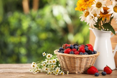 Wicker bowl with different fresh ripe berries and beautiful flowers on wooden table outdoors, space for text