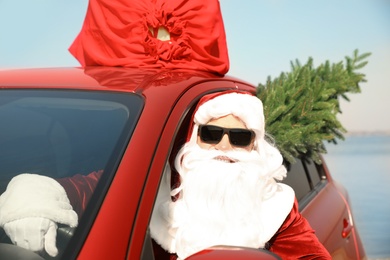 Photo of Authentic Santa Claus with fir tree and bag full of presents on roof driving modern car  near sea