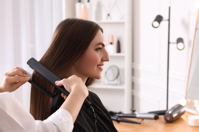 Photo of Hairdresser straightening woman's hair with flat iron in salon, closeup