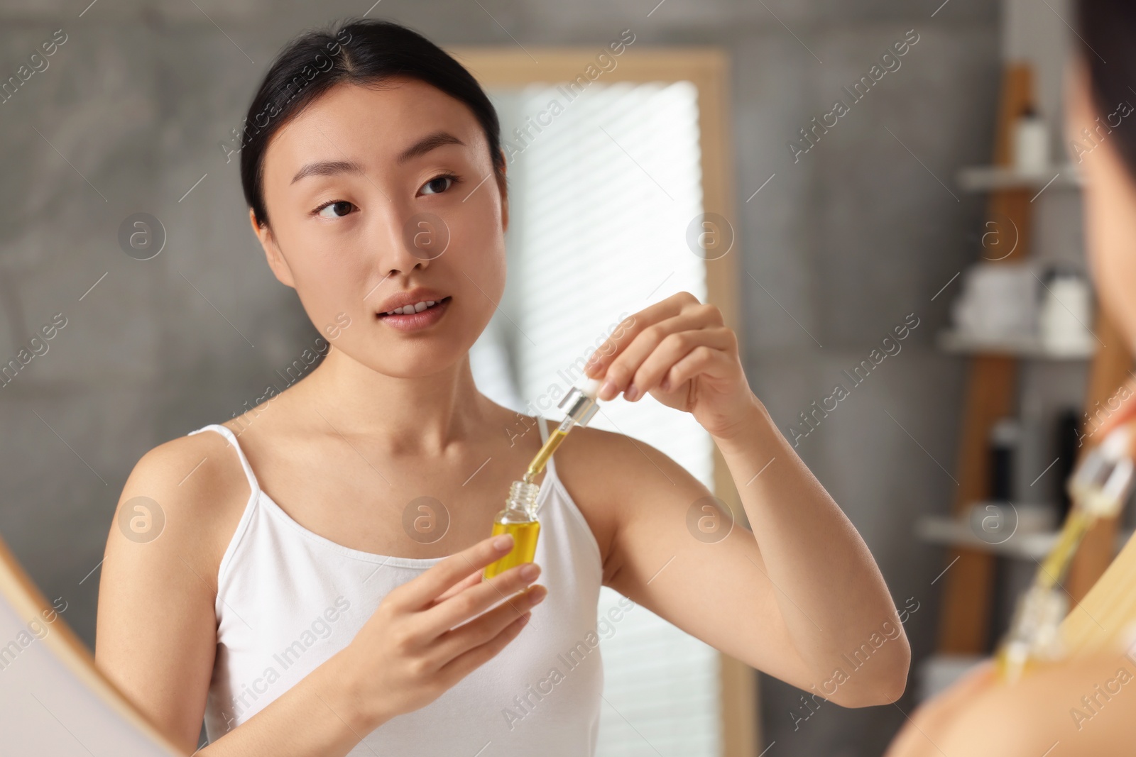 Photo of Beautiful young woman with bottle of cosmetic serum near mirror in bathroom