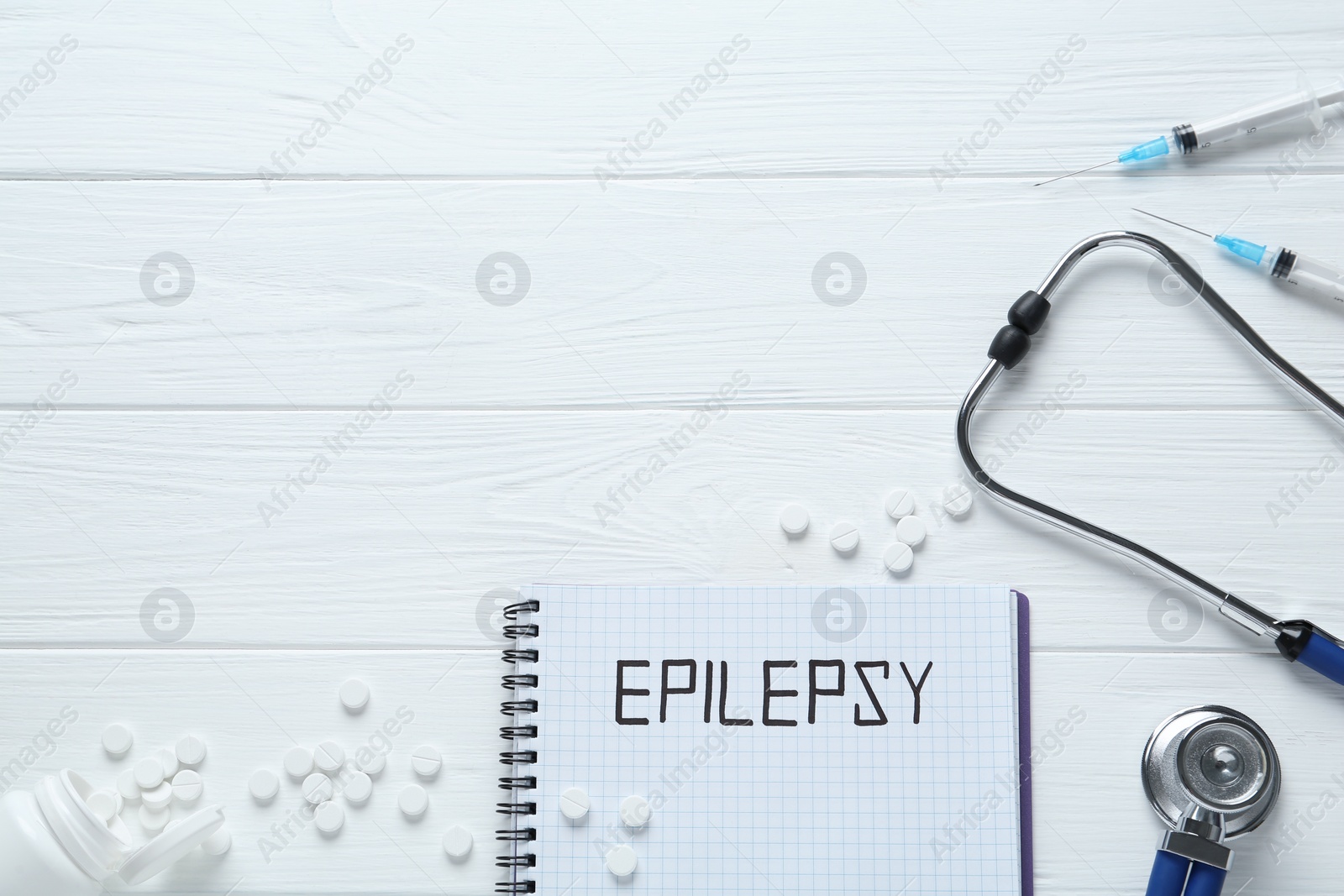 Photo of Notebook with word Epilepsy, stethoscope, pills and syringes on white wooden table, flat lay. Space for text